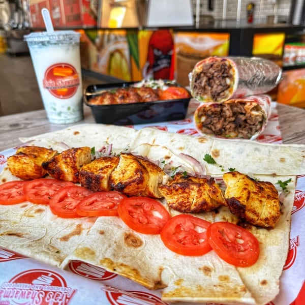Ara's Shawarma spread with chicken skewers, sliced tomatoes, shawarma wraps, and a drink.
