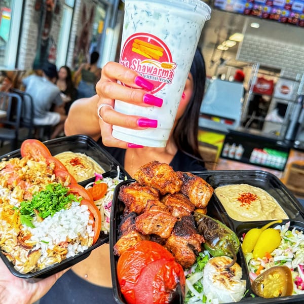 A person holds up two Ara's Shawarma takeout containers and a branded drink in a restaurant.