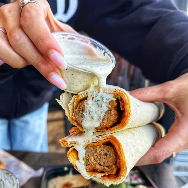 Hands pouring creamy white sauce over two halves of a delicious Ara's Shawarma wrap.