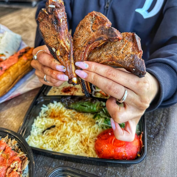 Hands holding two grilled lamb chops over a takeout container with rice, roasted pepper, and greens.