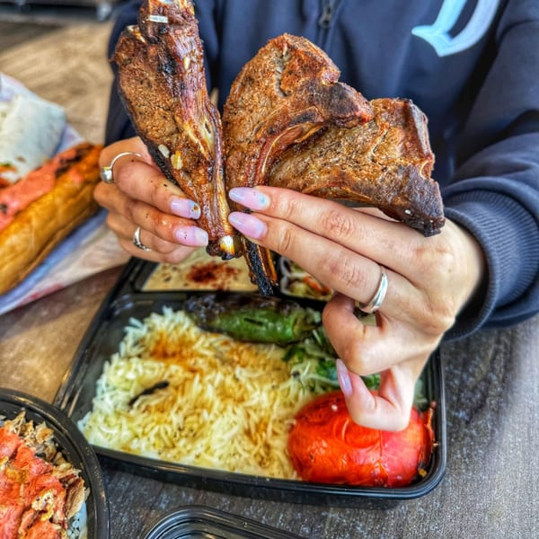 Hands holding two grilled lamb chops over a takeout container of rice, roasted pepper, and green pepper.